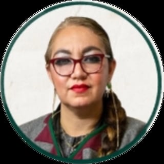 Portrait of a person with short blonde hair, burgundy-framed glasses, red lipstick, wearing a colorful striped shirt and green necklace