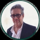 Middle-aged man with gray hair and dark-framed glasses wearing a dark blazer and white shirt, circular portrait photo