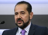Man with dark hair and beard wearing navy suit and purple tie speaking at microphone